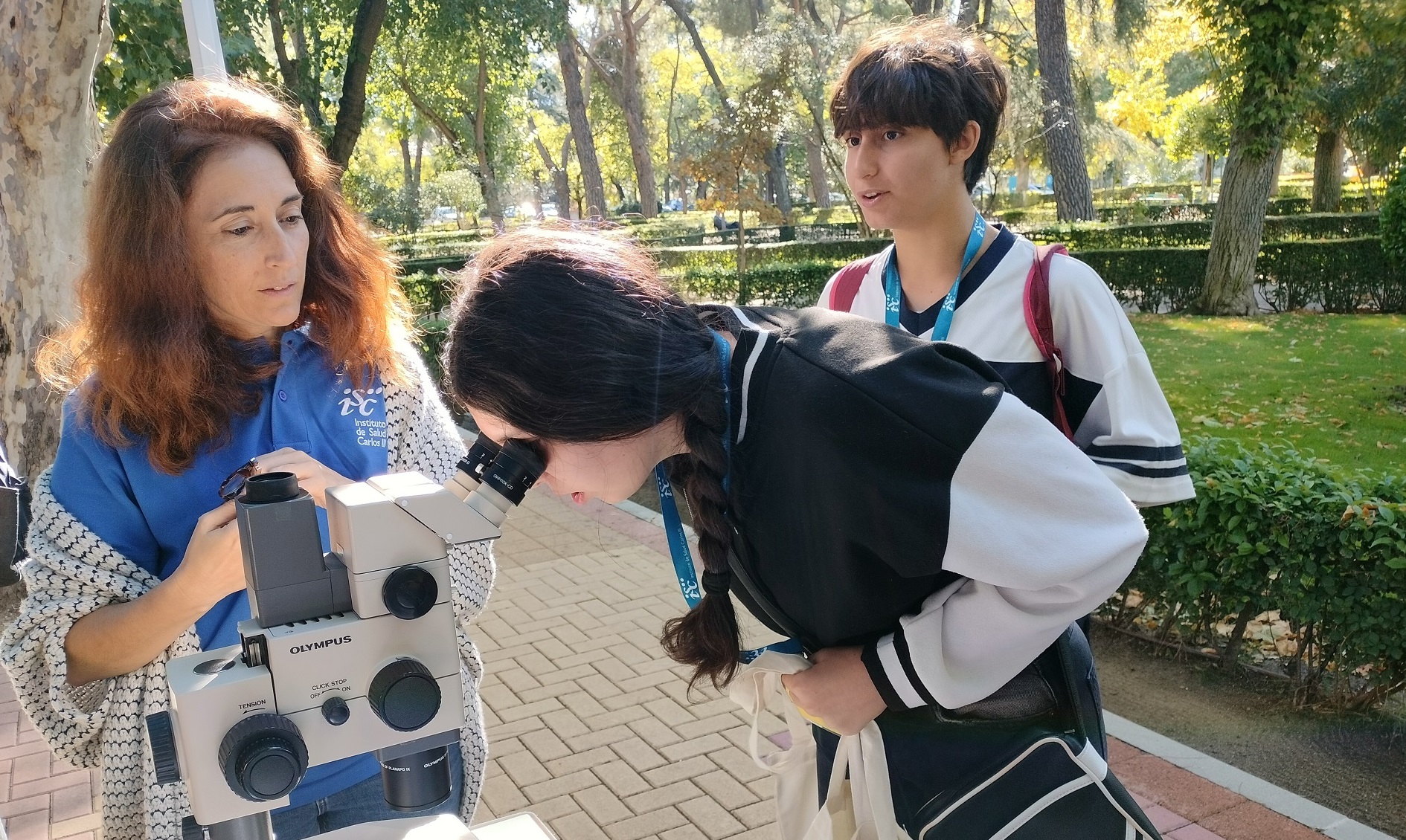 Una de las alumnas de los grupos de educación secundaria que visitaron el circuito científico del ISCIII mira una muestra en el microscopio, acompañada de una de las investigadoras del Instituto y de un compañero.